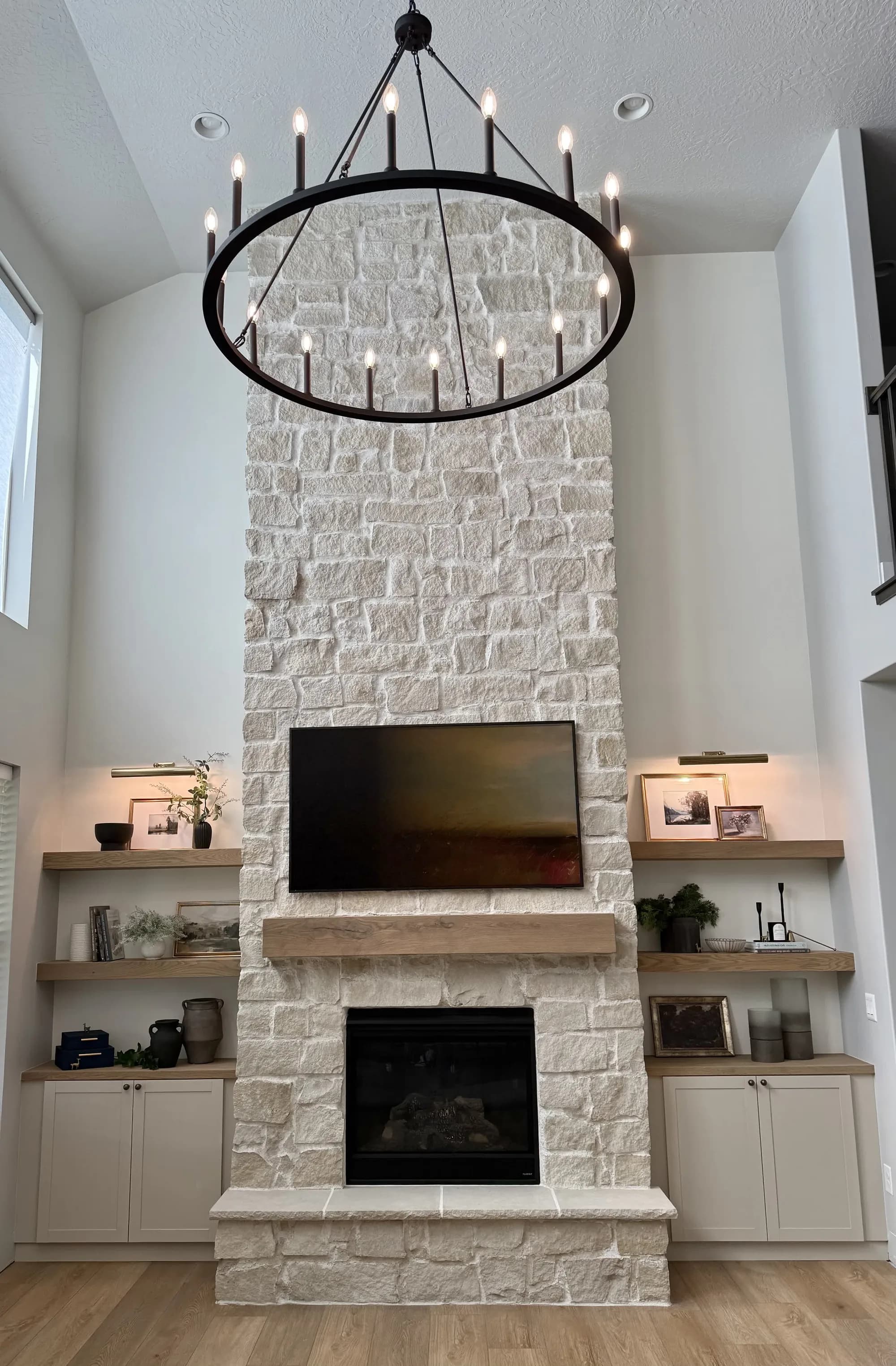 Two-story stone fireplace with floating wood shelves and chandelier by BK Toolbox